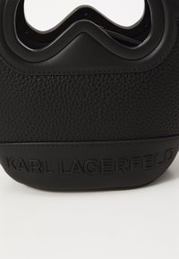 Czarna torba z teksturowanej skóry, z asymetrycznym otwarciem u góry i wytłoczonym brandingiem "KARL LAGERFELD" na dole.