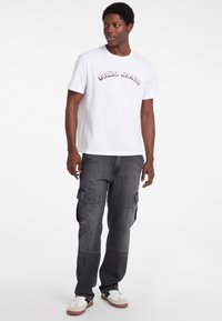 Tricou alb din bumbac cu textul "GUESS JEANS" în roz, asortați cu pantaloni cargo gri, cu buzunare laterale și o croială lejeră. Adidași maro și albi.