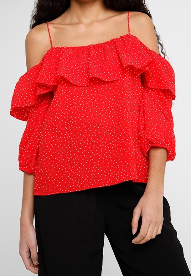 Femme portant un chemisier rouge à épaules dénudées avec des pois blancs et des volants, associé à un pantalon noir, montré des épaules aux hanches.