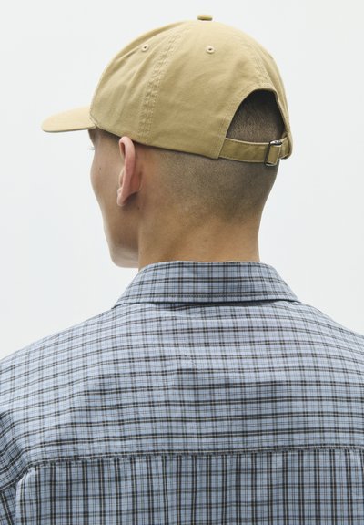 Lyhythiuksinen mies, jolla on beige baseball-lippalakki takaperin ja harmaa ruudullinen paita, nähtynä takaa yksiväristä taustaa vasten.