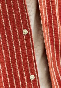 Close-up af et rødt tekstureret stof med cremefarvede lodrette striber og hvide knapper over en beige strikket beklædning.