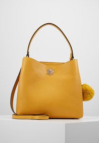 Bolso tote de piel amarilla con acabado texturizado, que presenta un cierre metálico redondeado y un adorno de pom-pom amarillo en un lado. Correa ajustable incluida.