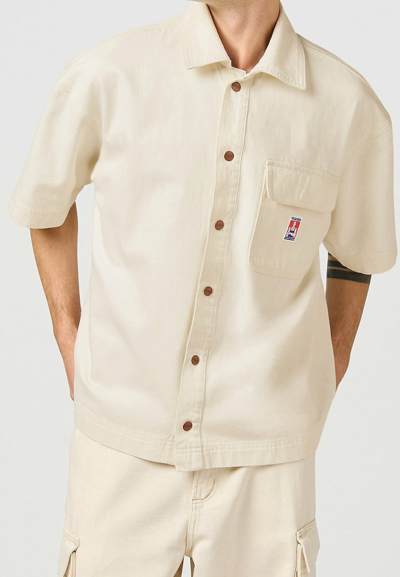 Camisa de manga corta color beige hecha de algodón, con cierre de botones en la parte frontal, un bolsillo en el pecho y etiqueta de marca. Presenta botones marrones y un corte relajado.