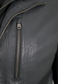 Schwarze Lederjacke mit einem silbernen Reißverschluss an der Brusttasche, mit glatter Textur und minimalistischer Gestaltung.
