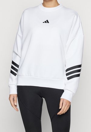 Osoba nosi białą bluzę z czarnym logo Adidas na piersi oraz trzema czarnymi paskami na każdym rękawie, w połączeniu z czarnymi legginsami.