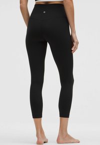 Leggings negros con una cintura alta, textura suave y diseño ajustado, que cuentan con un pequeño logotipo en la parte posterior. Pies desnudos visibles.