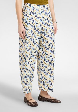 Lockere weiße Hose mit abstrakten blauen und braunen Blattmustern, getragen mit braunen gewebten Ballerinas und einem olivgrünen Oberteil.