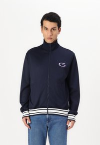 Giacca a zip blu navy con colletto alto, dettagli a righe bianche sui polsini e sull'orlo, caratterizzata da un piccolo logo sul petto.