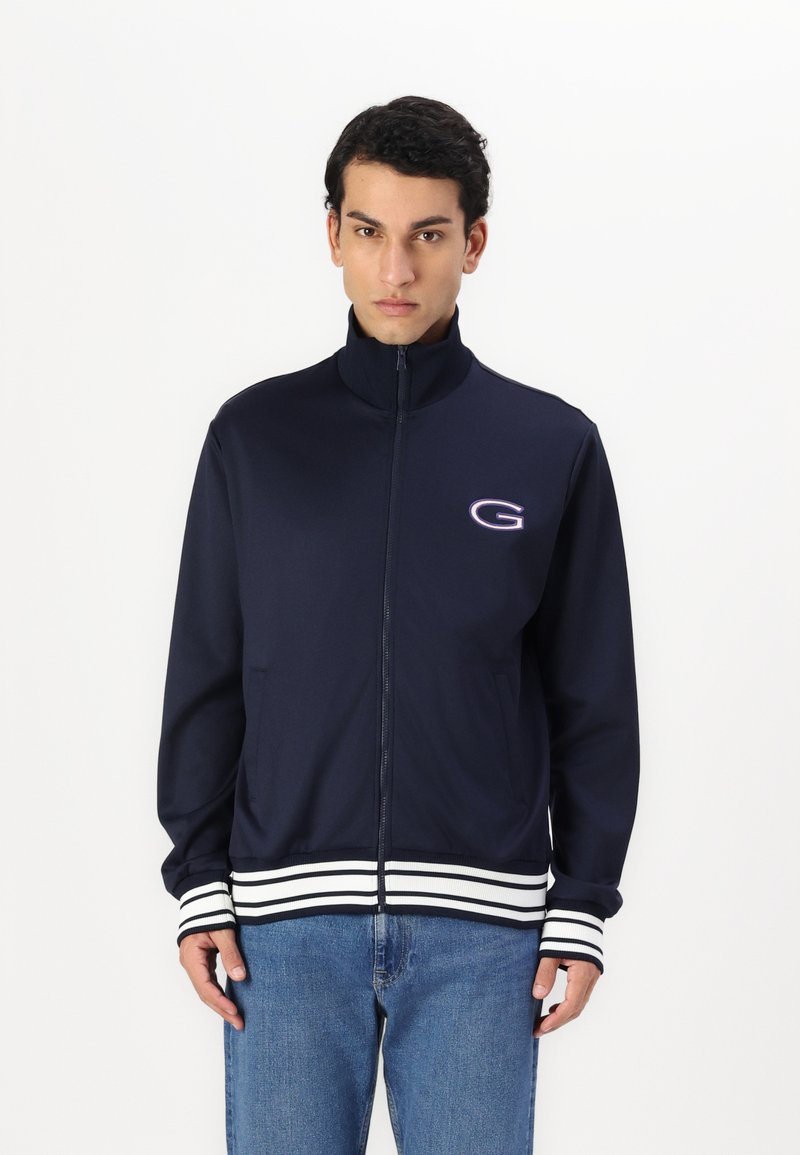 Giacca a zip blu navy con colletto alto, dettagli a righe bianche sui polsini e sull'orlo, caratterizzata da un piccolo logo sul petto.