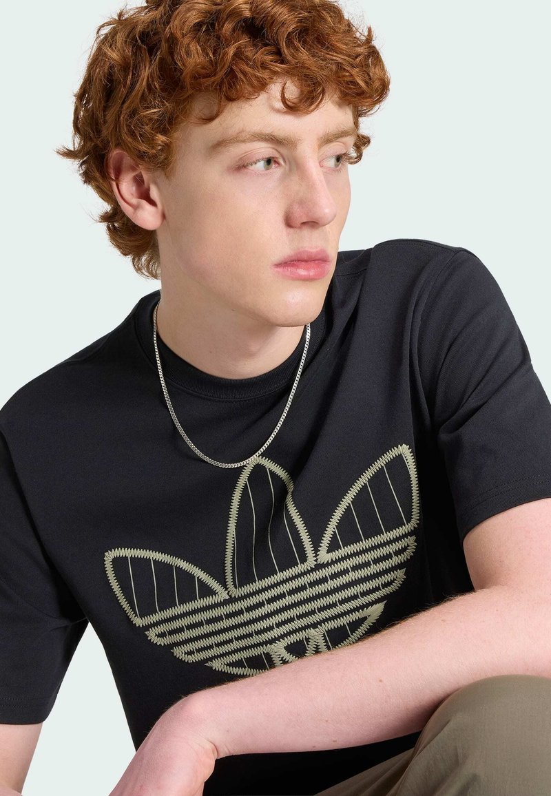 Jeune homme aux cheveux roux bouclés portant un t-shirt noir avec un grand logo beige de trèfle, chaîne en argent, regardant sur le côté sur un fond pâle.