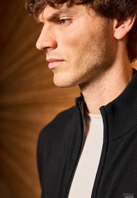 Černá zipová bunda s žebrovaným límcem, vyznačující se hladkým povrchem. Nositá přes bílou košili, ukazující detailní boční profil.