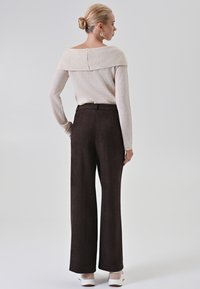 Maglione in maglia avorio con collo off-shoulder, abbinato a pantaloni larghi in tessuto marrone scuro. Modello mostrato di spalle, con sneakers.