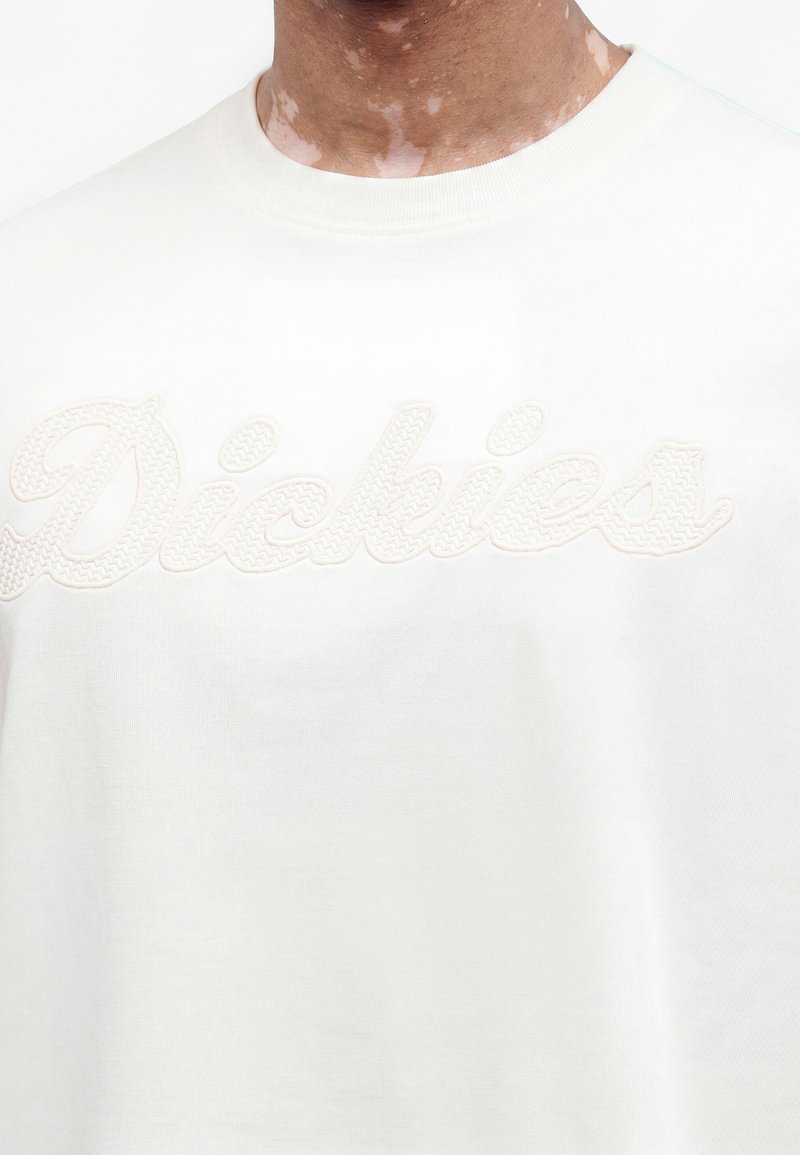 Nahaufnahme von Hals und Brust, die einen weißen Sweatshirt mit erhabenem, texturiertem "Dickies"-Logo zeigt, das über die Brust gestickt ist.