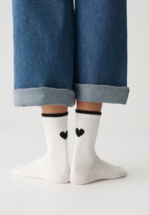 Weiße gerippte Socken mit schwarzen Bündchen, die kleine schwarze Herzdesigns auf der Rückseite haben, getragen mit hochgekrempelten blauen Jeans.