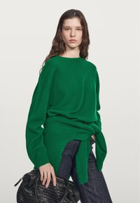 Grüner Strickpullover mit lockerem Sitz, langen Ärmeln und rundem Ausschnitt. Kombiniert mit dunklen Denim-Jeans und einer strukturierten schwarzen Handtasche.