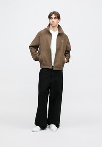 Hnědá manšestrová bunda s vysokým límcem, předními kapsami a elastickými manžetami, v kombinaci s volnými černými kalhotami a bílými teniskami.