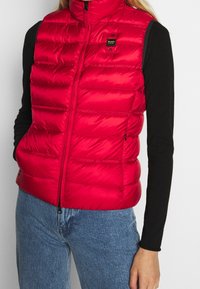Chaleco acolchado rojo con diseño de costura horizontal, cremallera en la parte delantera, cuello alto y una pequeña insignia en el pecho. Textura ligera y suave.