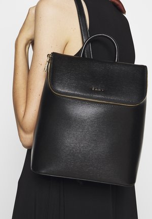 Zaino in pelle nera con texture liscia, cerniera dorata e manico superiore. Presenta un design minimalista con una tasca frontale.