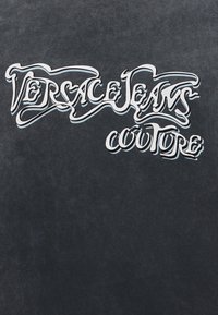 Λογότυπο Versace Jeans Couture με στυλιζαρισμένα λευκά γράμματα και μπλε σκιές πάνω σε σκουρόγκρι υφασμάτινο φόντο.