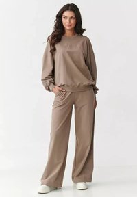 Brązowy, oversize'owy sweter z haftowanym napisem, w połączeniu z dopasowanymi spodniami typu wide-leg. Miękka tkanina z elastycznymi mankietami i wydobnioną talią. Białe buty.