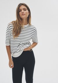 Bijeli i crni prugasti džemper s kratkim rukavima, okruglim izrezom i rebrastim rubom, uparen s pripijenim crnim hlačama. Jednostavan, ležeran dizajn.