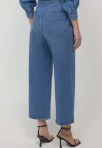 Jeans in denim blu a vita alta, con una vestibilità ampia e due tasche posteriori, abbinati a sandali neri con tacco alto. Tessuto testurizzato e leggero scolorimento.