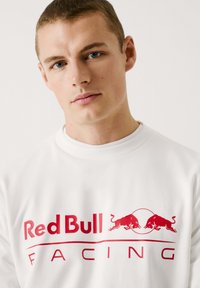 Bijela dukserica s okruglim izrezom, s velikim crvenim logom "Red Bull Racing" i dva crvena bika na prednjoj strani. Glatka tekstura.