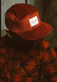 Gorra de pana roja con una etiqueta blanca que dice "Blackslides", con un visera estructurada. Llevada con una camisa de franela a cuadros rojos y negros.