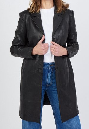 Person iført en sort læderfrakke over en hvid skjorte og blå jeans, holder frakkens revers med begge hænder.