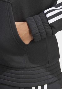 Sudadera negra con puños acanalados y un bolsillo lateral. Presenta rayas blancas en la parte superior de la manga. Diseño de tela suave y texturizada.