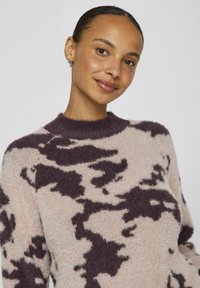 Pull en maille moelleuse rose clair avec un motif abstrait violet foncé, col rond et texture douce. Il présente des manches longues et une coupe décontractée.