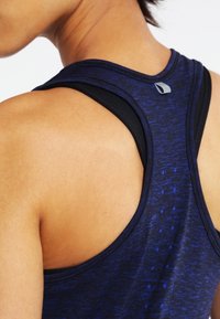 Námořnický modrý sportovní tílek s texturovaným vzorem, designem na zádech ve stylu racerback a malým šedým akcentem na výstřihu, který ukazuje prodyšný materiál.