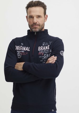 Felpa blu navy con colletto alto, chiusura con zip e motivi di testo ricamati in bianco e rosso. Presenta un pannello laterale e polsini a costine.