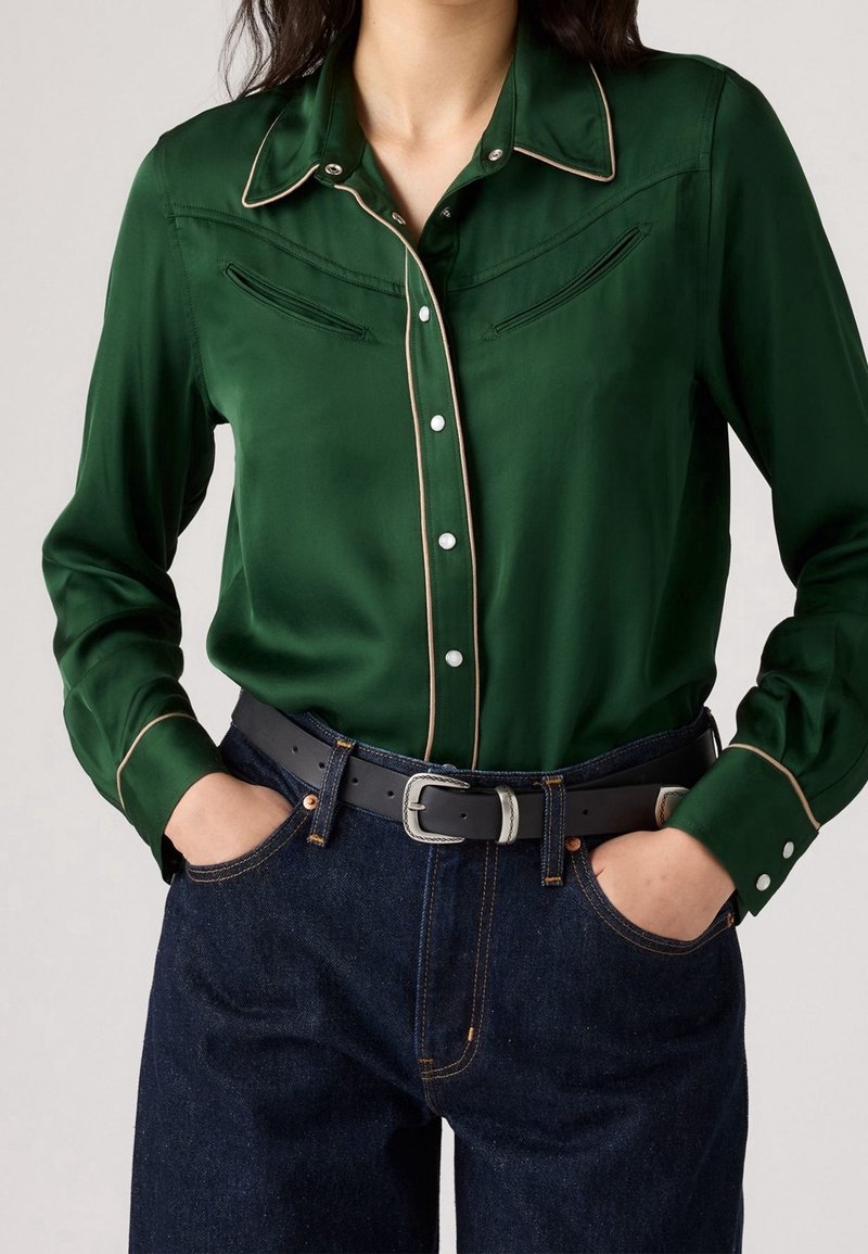 Femme portant une chemise style western satin vert foncé avec passepoil beige, jean bleu foncé, ceinture noire avec boucle argentée, mains dans les poches.