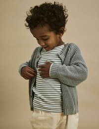 Grau gestrickte Strickjacke mit Knöpfen, getragen über einem gestreiften Hemd. Weicher Stoff, lässige Passform. Hellbeige Hosen vervollständigen das Outfit.