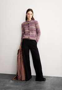 Une femme se tient contre un mur blanc, portant un cardigan à rayures mauve-brun et un pantalon noir à jambes larges, tenant une veste rayée marron dans sa main gauche.