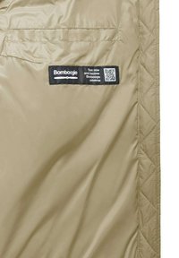 Tessuto trapuntato beige con un'etichetta nera con logo e codice QR. Texture liscia, evidenziando le cuciture e una tasca laterale.