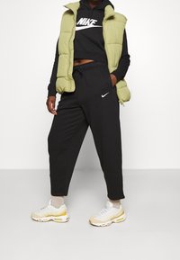 Grön pufferväst över en svart kort hoodie, kombinerad med svarta joggers och beige sneakers med gula accenter. Nike-logotyper synliga.