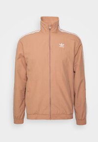Gaiši brūns zip-up jakas, ribināts apakšmalā un aproces. Iezīmētas ar baltām sānu svītrām un izšūtu logotipu uz krūtīm. Gluda auduma tekstūra.