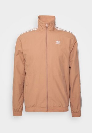 Šviesiai rudas zip-up džemperis, lanjutis apdaila ir rankogaliai. Savybėmis balti šoniniai dryžiai ir išsiuvinėtas logotipas ant krūtinės. Lygus audinio paviršius.