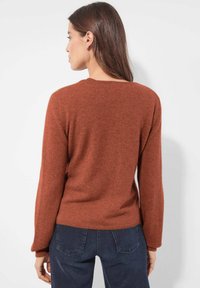 Vrouw met lang bruin haar, gekleed in een rustkleurige trui en donkerblauwe jeans, met haar rug naar de camera gekeerd tegen een effen witte achtergrond.