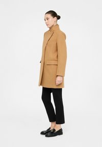 Cappotto in misto lana color cammello con colletto alto, zip frontale e tasche laterali. Indossato su pantaloni neri aderenti e mocassini neri. Tessuto liscio.