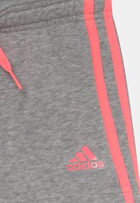 Pantalones deportivos grises con rayas rosas y cordón ajustable. Presentan un logo de Adidas en rosa en la parte baja de la pierna. Textura de tela suave.