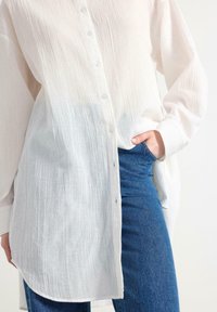 Camisa blanca con textura y un degradado; de manga larga, corte holgado, bajo ligeramente curvado y pequeños botones transparentes.