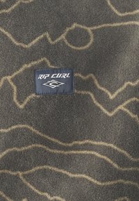 Fleecowy materiał Rip Curl w ciemnoszarym kolorze z brązowym falistym wzorem. Zawiera granatową metkę marki z białym napisem. Miękki, teksturowany materiał.