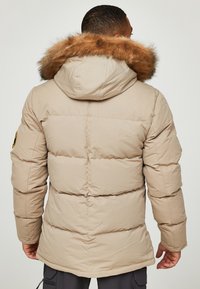 Chaqueta de plumas beige con capucha forrada de piel, que presenta un diseño acolchado y puños elásticos. Incluye un parche de logo en la manga izquierda.