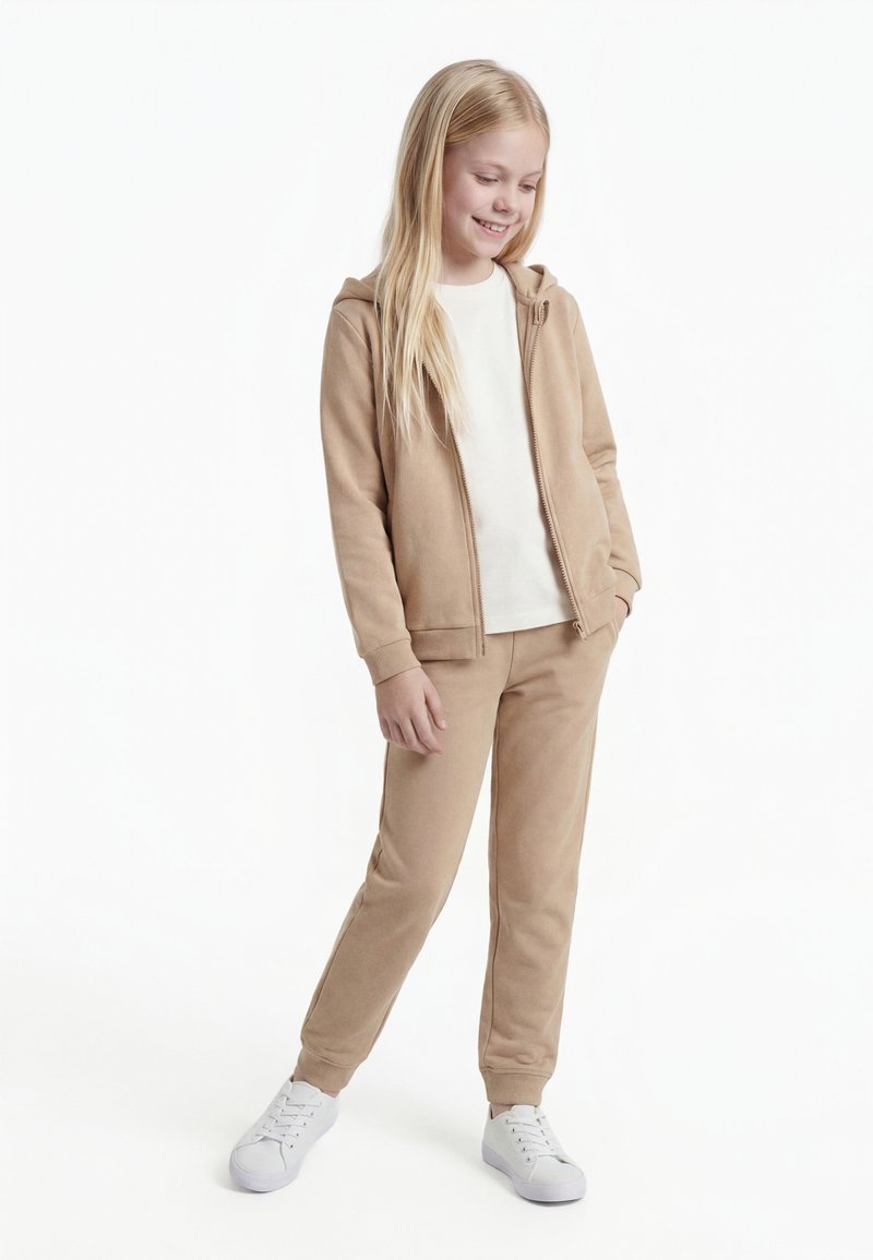 Jauna mergaitė su ilgais blondiniais plaukais šypsosi, vilki smėlio spalvos megztinį su kelnėmis ir baltus sportbačius, stovi ant baltos fono.