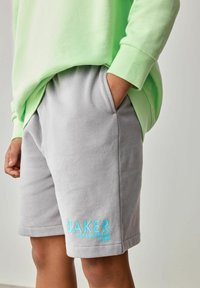 Grijze katoenen shorts met zijzakken, voorzien van turkoois geborduurde tekst "BAKER OPGERICHT 1983," gecombineerd met een lichtgroene sweatshirt.