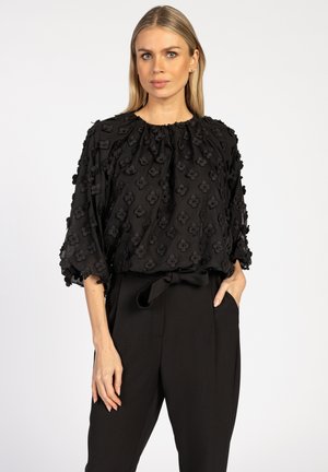 Zwarte blouse met een gestructureerd bloemenpatroon, korte puffmouwen en een geplooide halslijn, gecombineerd met hoge zwarte broeken.