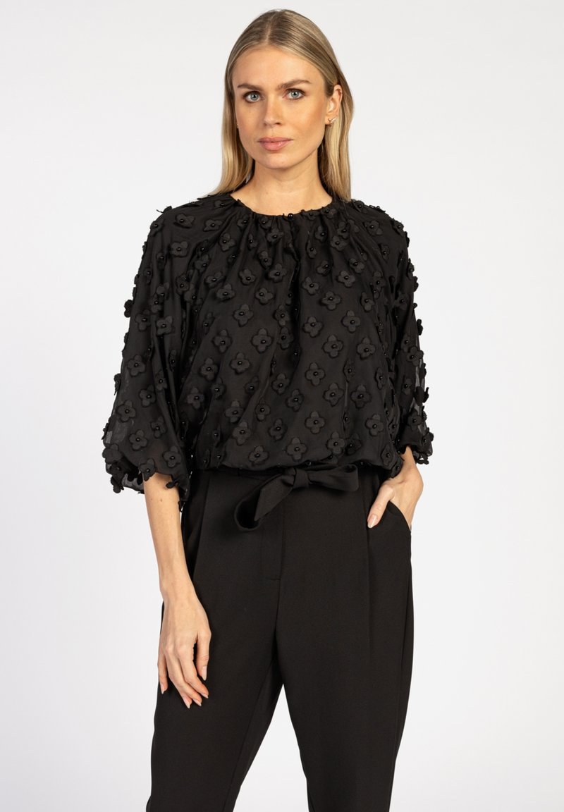 Zwarte blouse met een gestructureerd bloemenpatroon, korte puffmouwen en een geplooide halslijn, gecombineerd met hoge zwarte broeken.